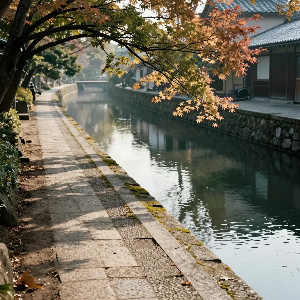 Quiet Kyoto canal path with stone edging, maples, mist, and a temple roof beyond water.